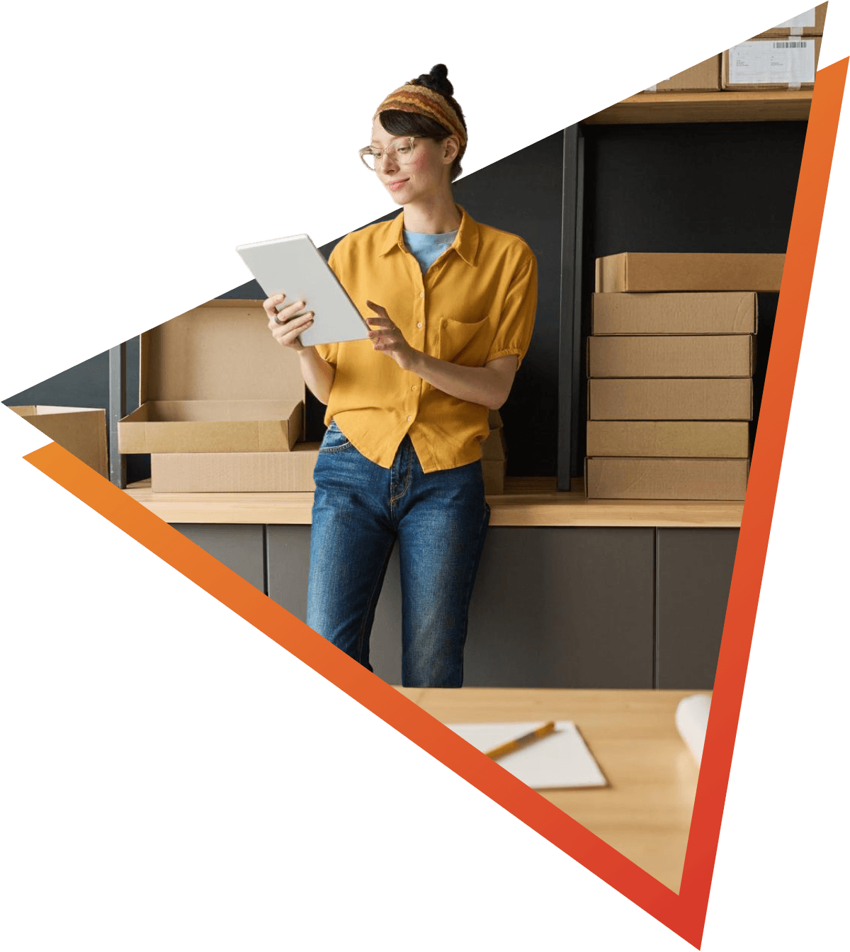 Young woman in casual clothing holding a tablet in a work setting. Around her, there is a desk, packing boxes, and a notebook.